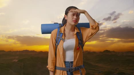 excursionista asiática con mochila de montaña mano frente sonriendo y mirando la distancia mientras está de pie en la cima de la montaña durante el atardecer