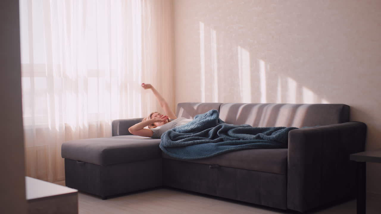 Freelancer stretches after sleep on sofa in living room, covered with blanket, side stool close to couch, warm sunlight shining through curtains, peaceful cozy home