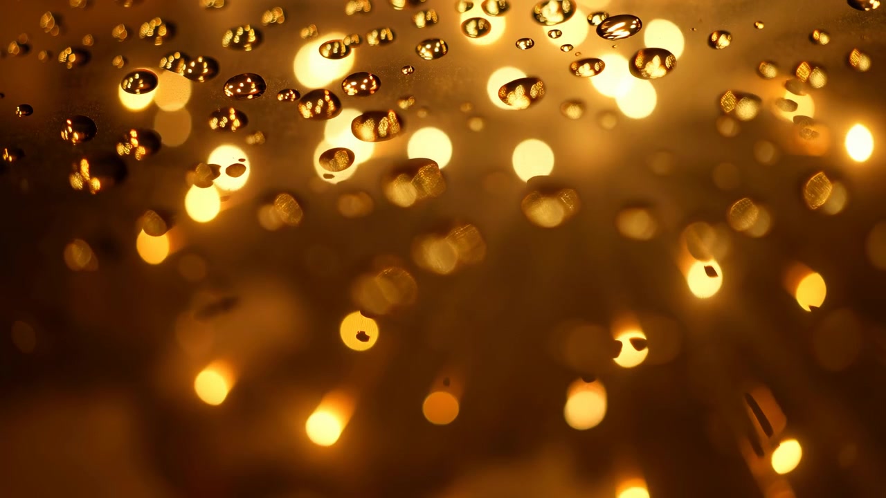 Vista ravvicinata di goccioline d'acqua limpide su vetro inclinato, con dense luci ambrate calde dietro, riflessi luminosi, sfocatura soffusa e striature radiose che formano una ricca texture astratta, con goccioline d'acqua sul vetro.