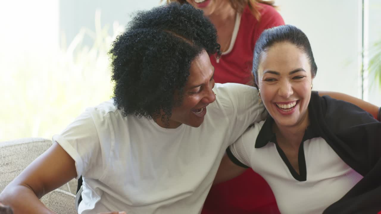Diverse friends left man initiating hug, laughing, shifting on couch by bright window and plant