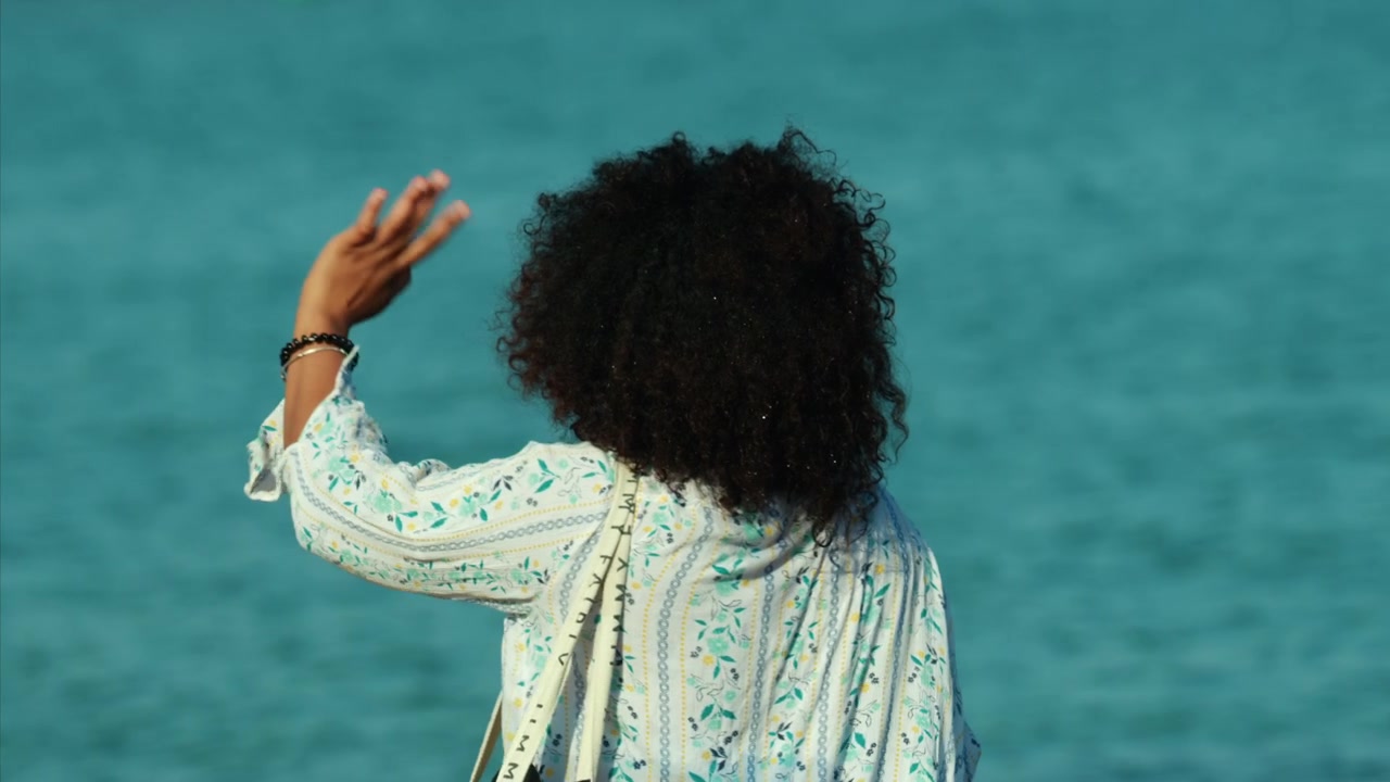 Woman with afro hairstyle stands alone, looking at the vibrant turquoise-blue ocean. Solitude and freedom concept