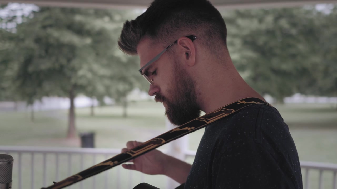 Uomo che suona la chitarra e canta sotto un gazebo sotto la pioggia - Inquadratura laterale che si sposta sul retro
