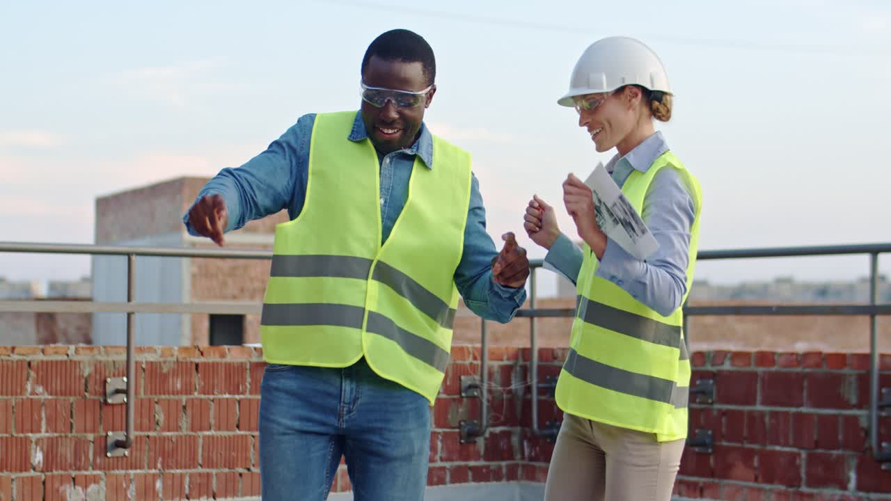 Young and attractive African American cheerful man and happy Caucasian woman, builders in hardhats dancing on the roof of the building at the construction site. Outside.