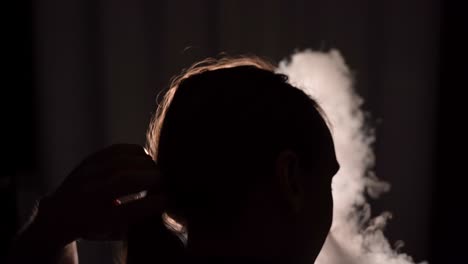 isolated silhouetted closeup of a man untying his long hair while binge smoking in a dark room facing a wall