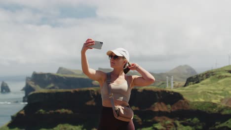 chica de viaje con gafas de sol tomando selfies frente a la costa del océano y acantilados verdes durante un día de viento