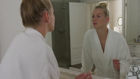 Caucasian-woman-in-bathrobe-touching-her-face-while-looking-in-the-mirror-in-the-bathroom