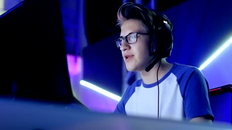 teenage boy gamer plays in competitive video game on a esports tournament/ internet cafe. he wears glasses and headphones and speaks into microphone.