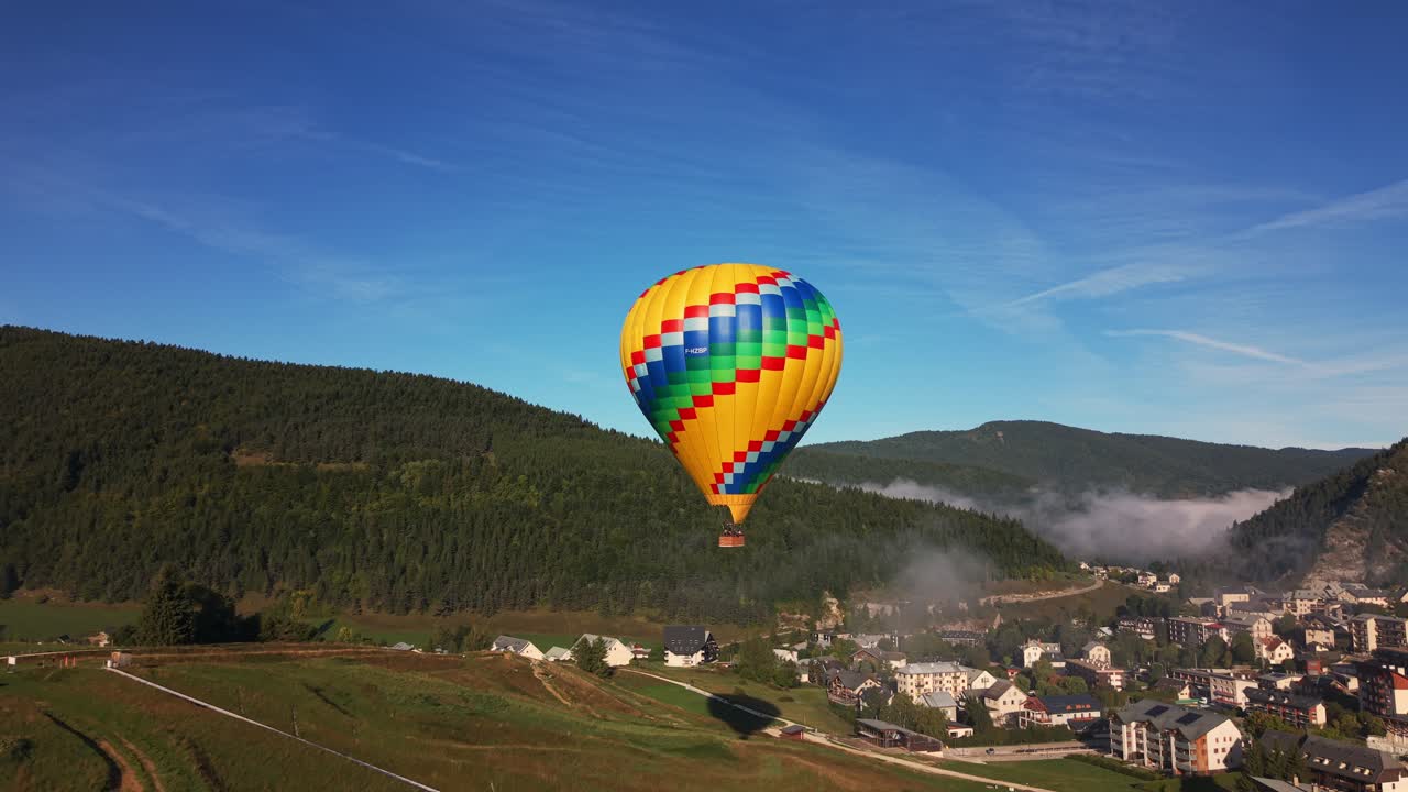 Um vibrante balão de ar quente flutua sobre uma pequena aldeia francesa, colinas enevoadas e uma paisagem de floresta verde.