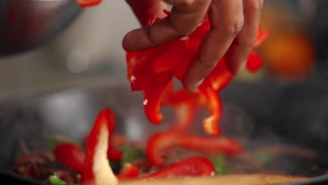 hand throwing chopped red bell peppers into a frying pan with cooked onions