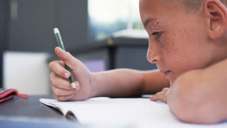 Un-Niño-Birracial-Con-Pecas-Escribe-En-Un-Cuaderno-En-Un-Salón-De-Clases