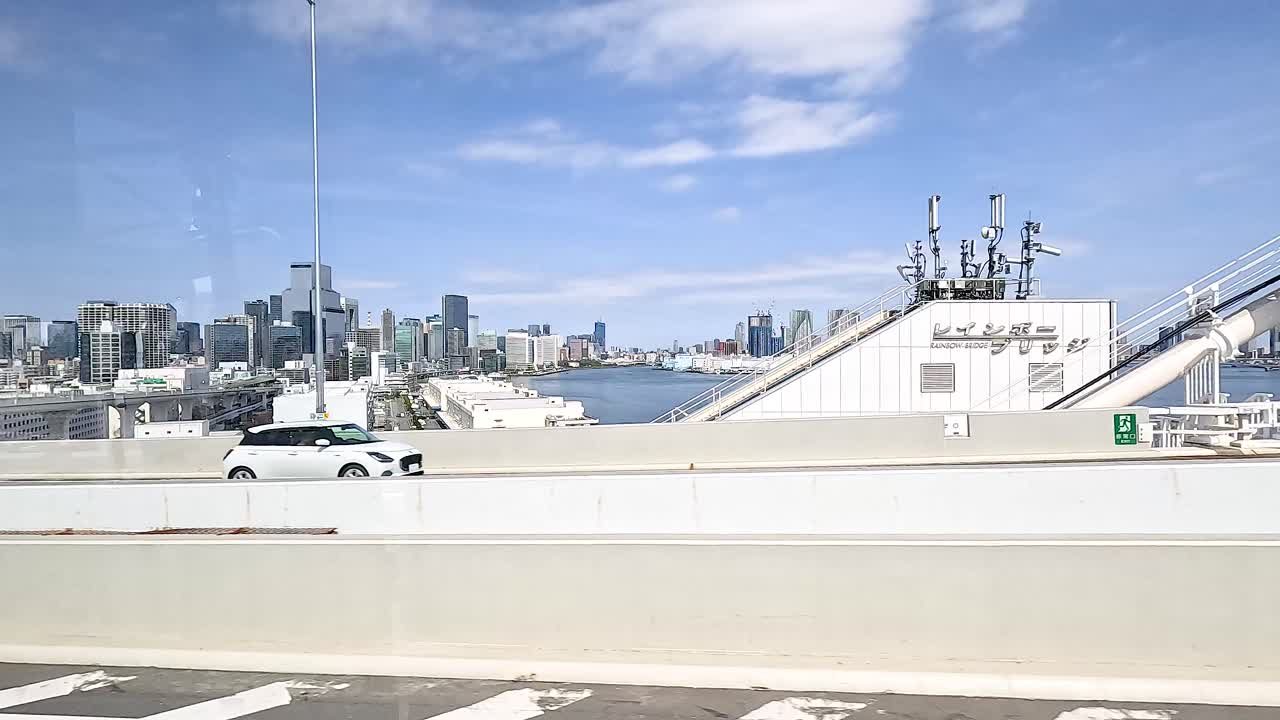 Vehicles traverse a Tokyo bridge under clear skies, with the city skyline in the background. Dynamic movement and urban scenery