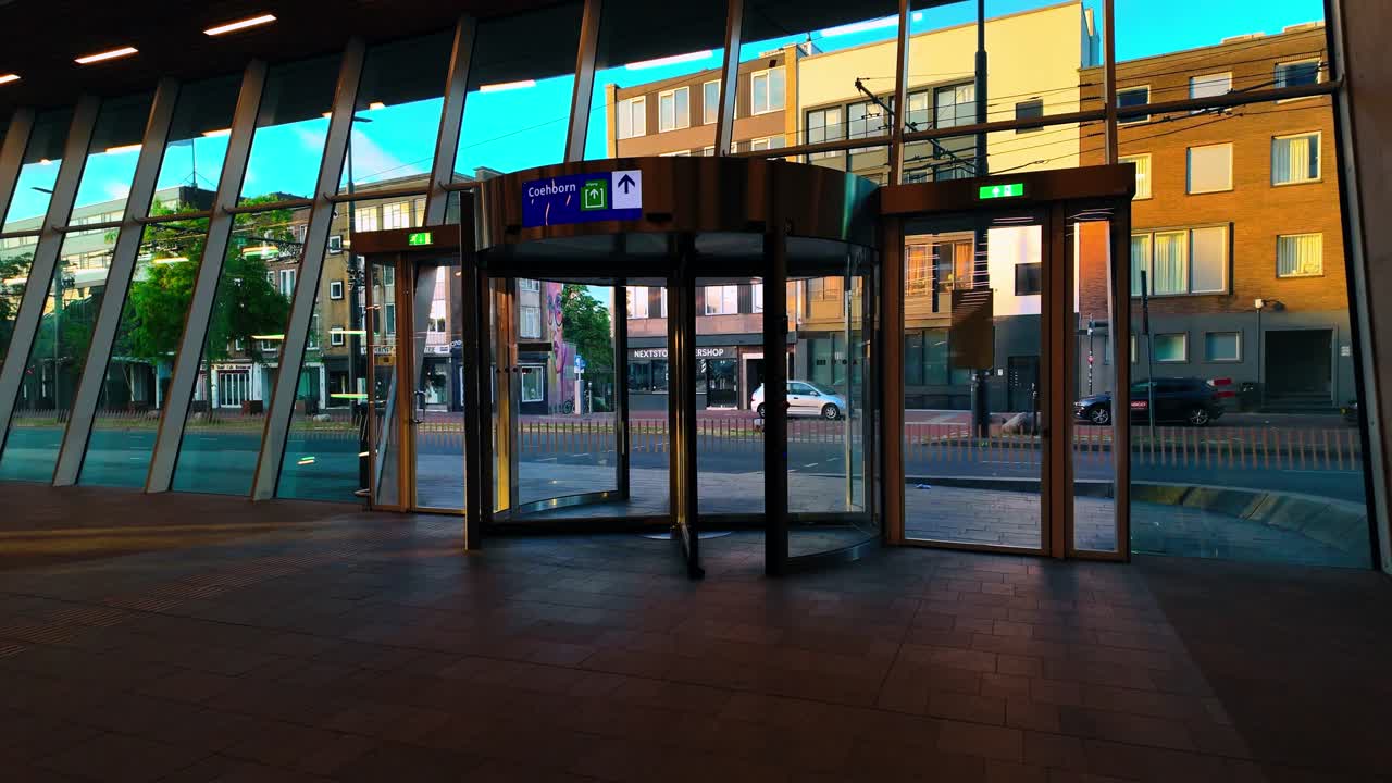 Modern interior view of revolving glass doors at Arnhem Central Station during golden hour sunset. Large windows reveal city street architecture and blue sky in Gelderland Netherlands