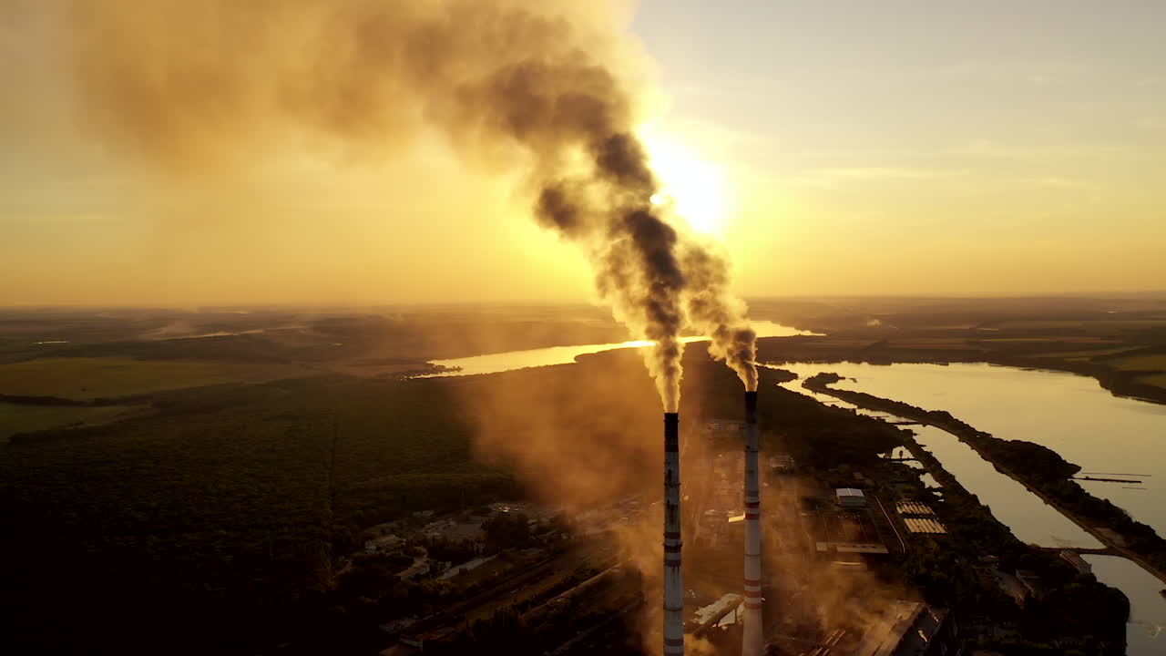 Dirty emissions coming out of industrial pipes. Factory surrounded by nature in the evening. Thick smoke going into th air from manufacturing at sunset. Pollution.