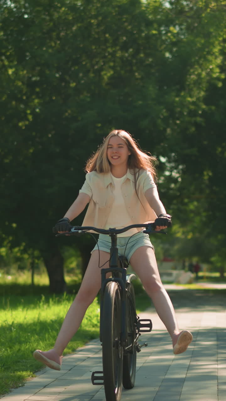 젊은 여성은 푸른 공원 길을 따라 자전거를 자신감 있게 타고, 균형을 위해 한 다리를 약간 펴고, 배경에는 활기찬 나무, 램프 기둥, 그리고 두 사람이 뒤에서 고 있습니다.