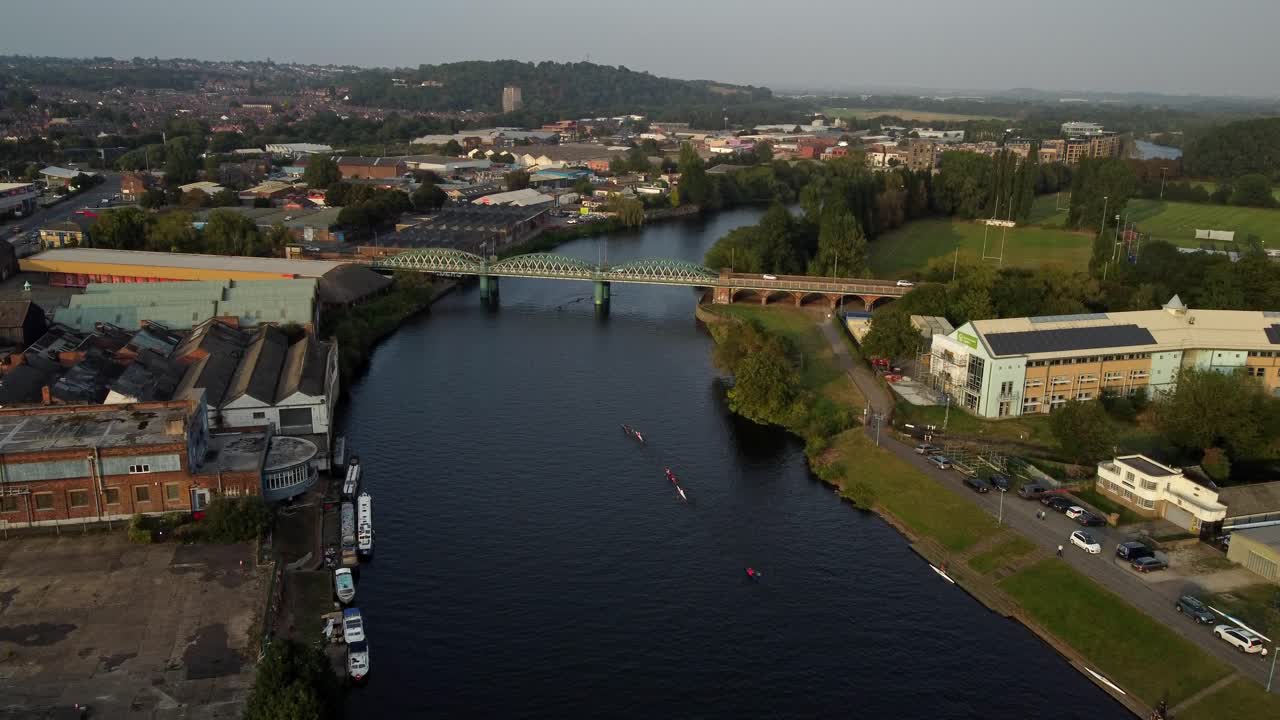 Video of three rowing boats on the River Trent in Nottingham City Centre heading towards a bridge. The footage starts high capturing the industrial and residential surroundings before lowing down.