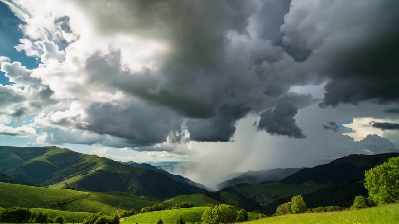 Büyüyen bir kümülonimbus bulutu, yeşil tepelerin üzerine yoğun bir yağmur sütunu bırakarak vadi yerleşimini gizliyor.