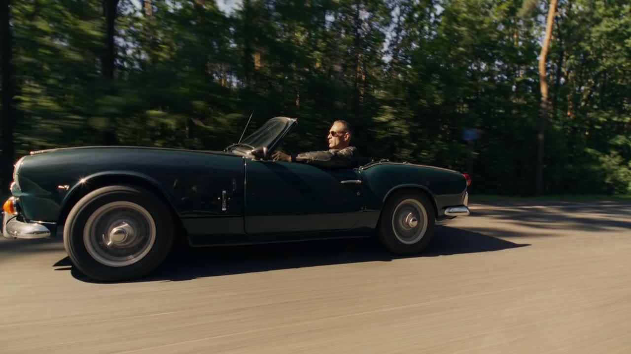 A classic car speeds down a tree-lined road on a sunny day