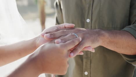 hands of diverse bride putting wedding ring on finger of groom at beach wedding, in slow motion