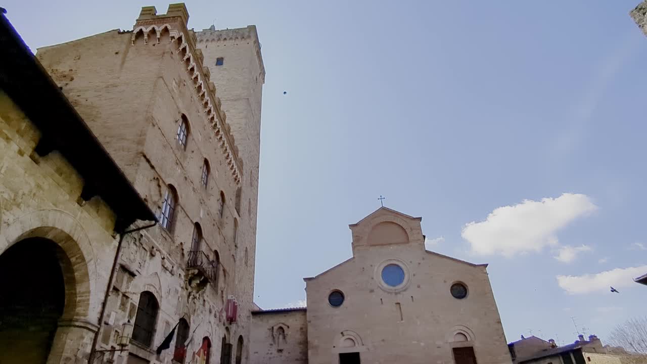 Footage of birds flying around the Duomo di Santa Maria Assunta. The Duomo is in San Gimignano, Italy. The footage shows the architecture of the church and the natural movement of the birds.