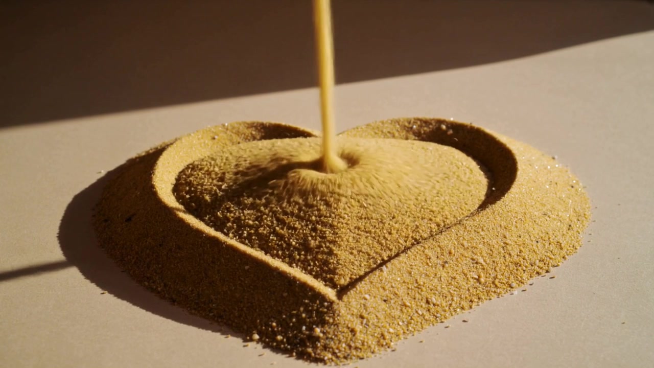 Narrow stream pouring fine golden sand shaping heart mound on white sheet, rim highlighted