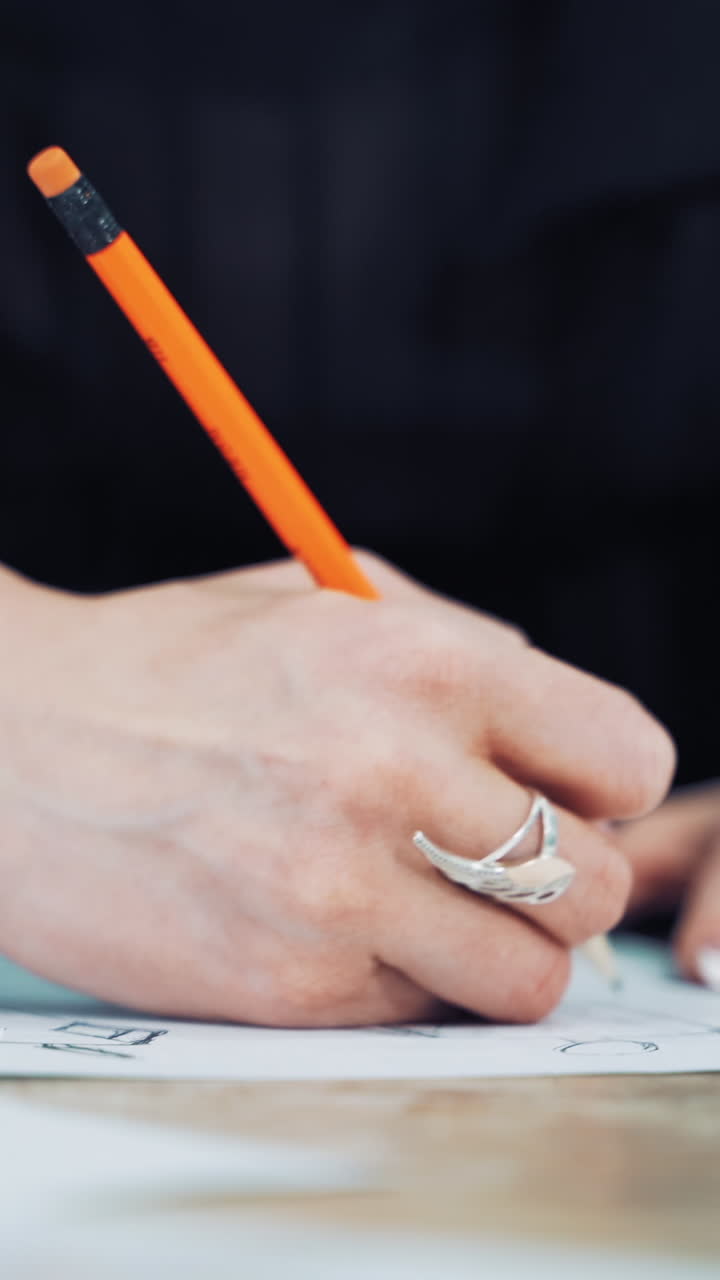 Hands of a professional tailor is drawing a sketch with a sharp pencil on a table. Dressmaker is making a creative design of a dress on a sheet of paper in her workplace. Vertical video