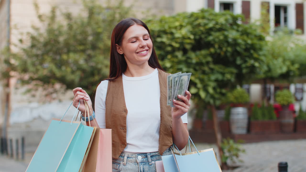 Happy young shopaholic caucasian woman holding cash while carrying shopping bags on city street
