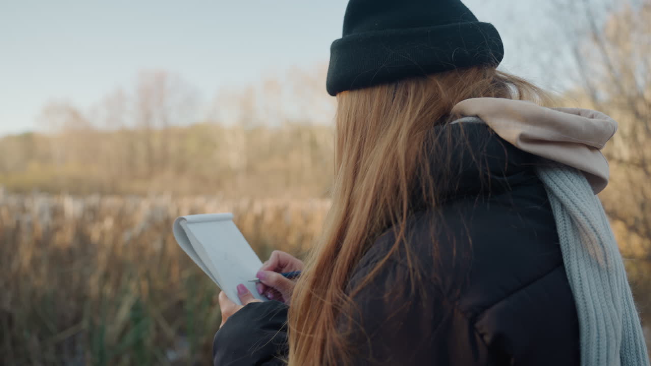 Rear view field notebook recording nature, woman in beanie and scarf with heavy coat, marshland reeds and wide horizon, focused pen movements and steady posture, calm sunset light and gentle breeze