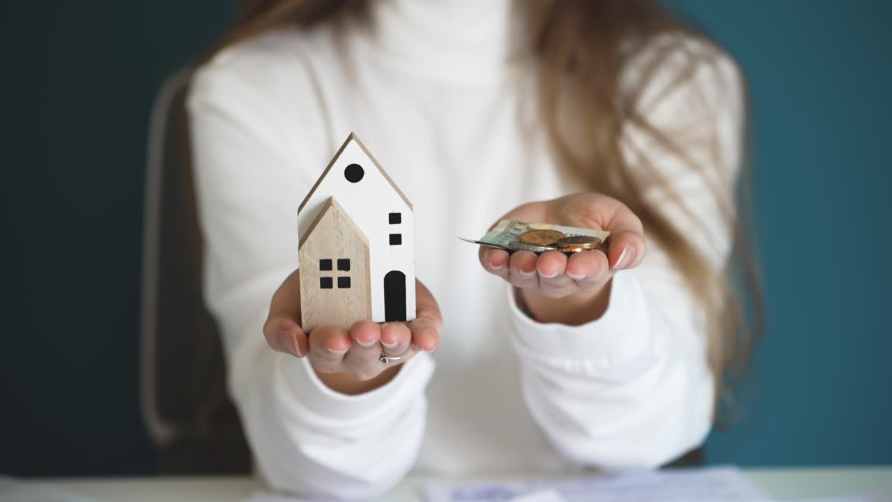 Donna che tiene contanti in una mano e una casa in miniatura nell'altra, illustrando la pianificazione finanziaria, il risparmio di denaro e la gestione del budget per l'acquisto di una nuova casa o un investimento immobiliare.