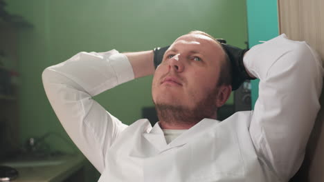 a close-up view of a technician in a white lab coat and black gloves, leaning back and relaxing with hands behind his head in a lab setting