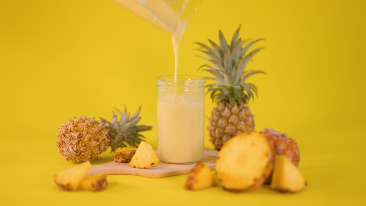Creamy pineapple smoothie poured into glass, surrounded by fresh fruit, on bright yellow background