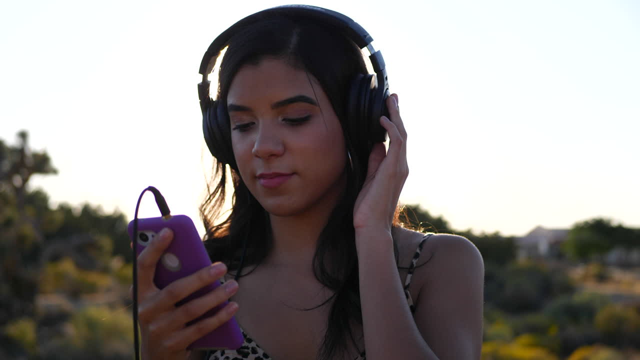 una hermosa joven hispana sosteniendo un teléfono inteligente escuchando música con auriculares al aire libre bajo la épica luz del sol con bengalas en cámara lenta