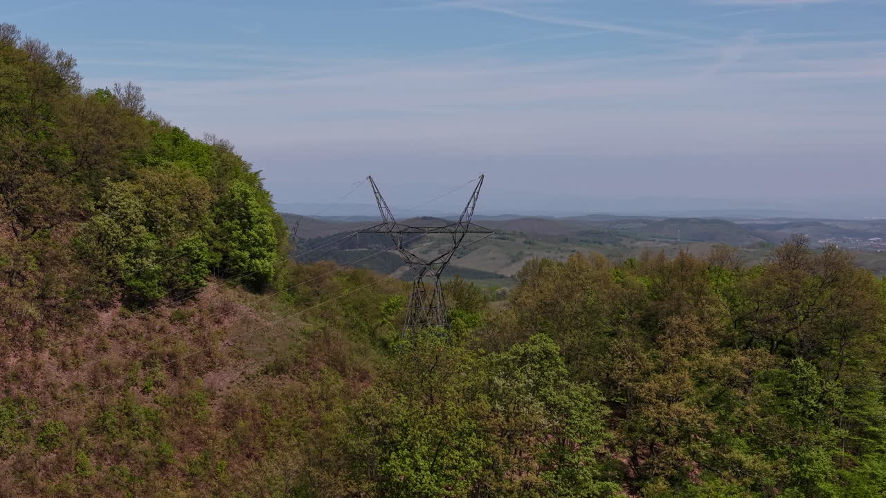 Vysokonapěťový stožár se výrazně tyčí na zalesněném svahu, shlíží na širokou venkovskou krajinu a vzdálené město pod modrou oblohou.