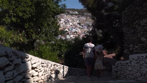 Back-view-of-couple-in-summer-outfit-walking-down-stone-steps-with-backdrop-of-beautiful-greek-village-on-hill