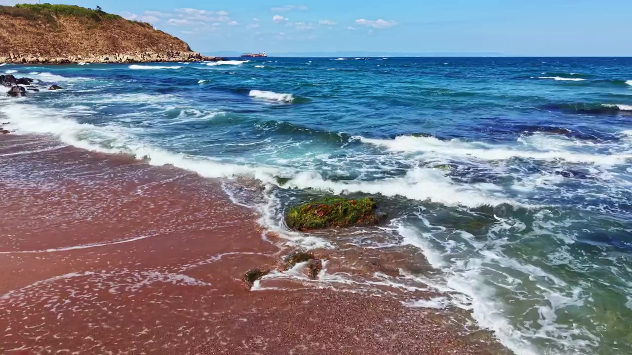 Onde si infrangono sulla riva di una spiaggia con scogliere rocciose sotto un cielo azzurro limpido nel pomeriggio
