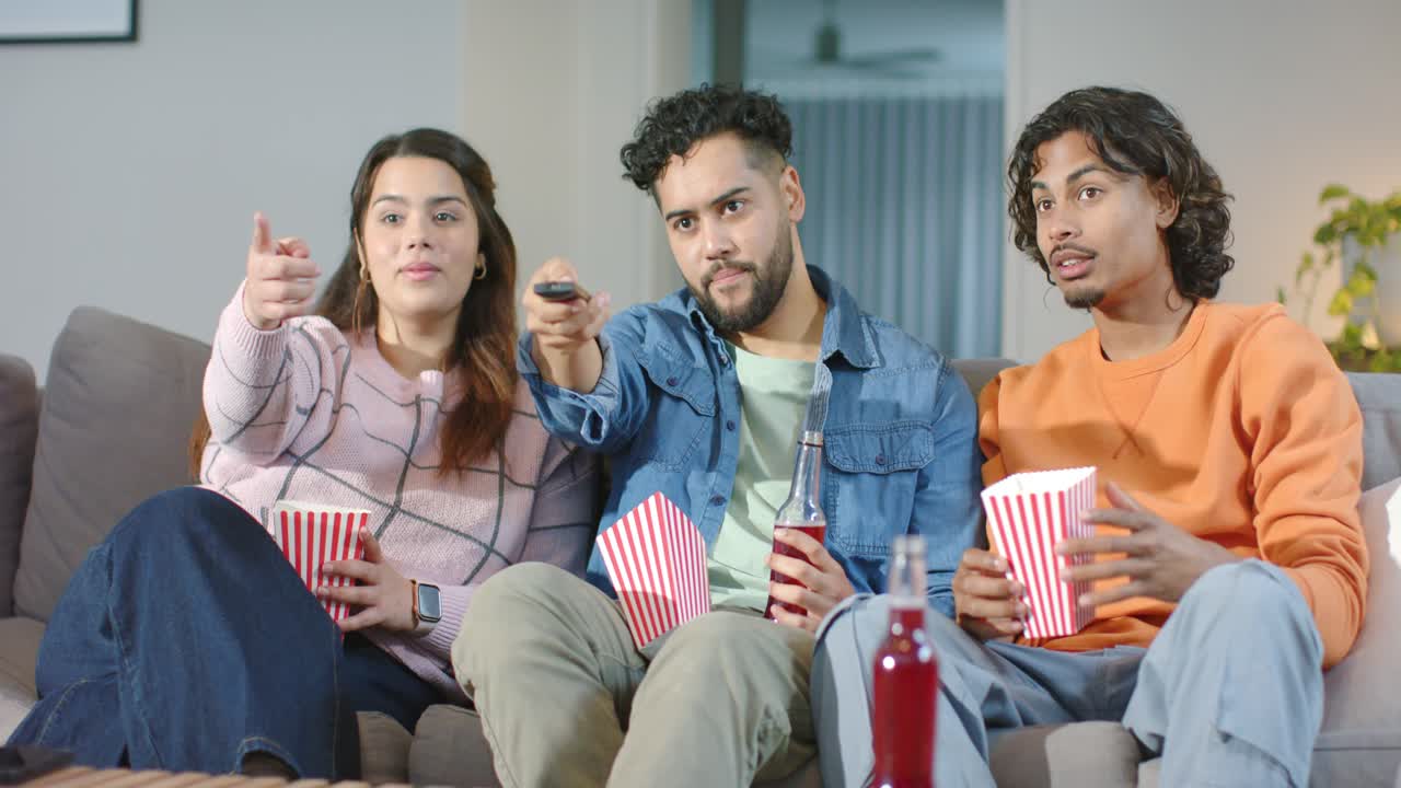 Gruppo di amici eterogenei seduti sul divano che premono il telecomando per avviare uno show, mangiando popcorn e indicando lo schermo della TV