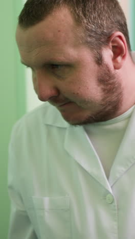 una vista cercana de un técnico de laboratorio en una bata blanca mirando hacia abajo, profundamente enfocado y buscando algo, con dos estantes detrás de él y la pared pintada de verde