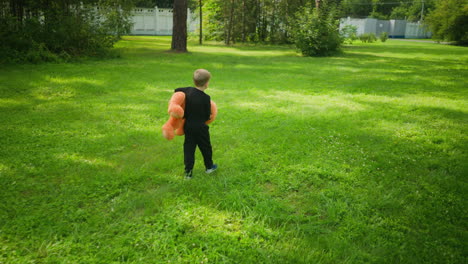 rear view of young kid dressed in black walking alone on green grassy field holding large orange teddy bear, with long shadows cast on ground and tall white fence visible in background under daylight