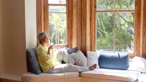 happy senior biracial woman sitting on couch and drinking tea at home, slow motion