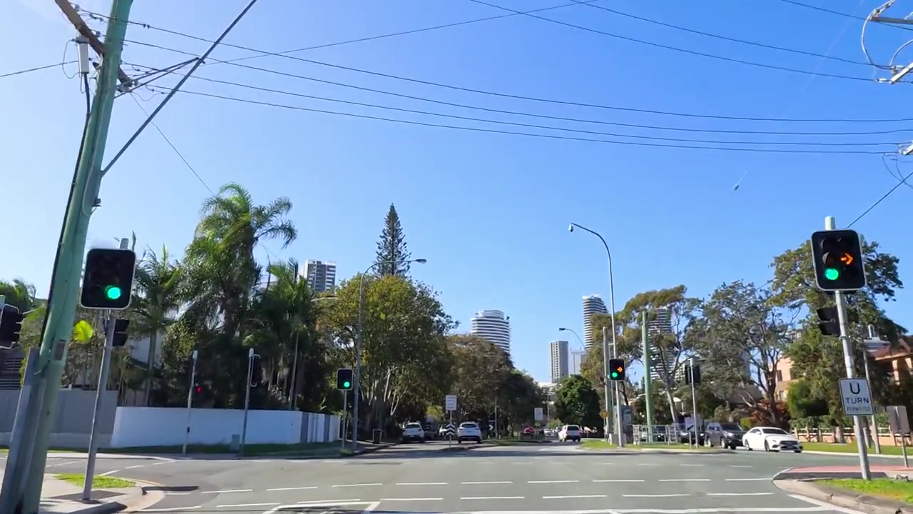 Vue d'un véhicule avançant le long d'une route de banlieue bordée d'arbres sous un ciel bleu clair, vers des gratte-ciel lointains.