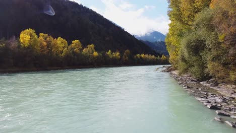 Flying-along-river-with-trees-growing-around,-mountains-in-the-distance-on-a-sunny-day