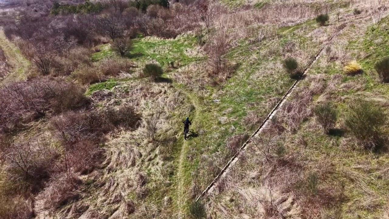 Gün ışığında, arka planda bir su kütlesinin yakınında, kuru bitkiler ve çalılıklarla çevrili dar bir patikada yürüyen doğa yürüyüşçüsü.