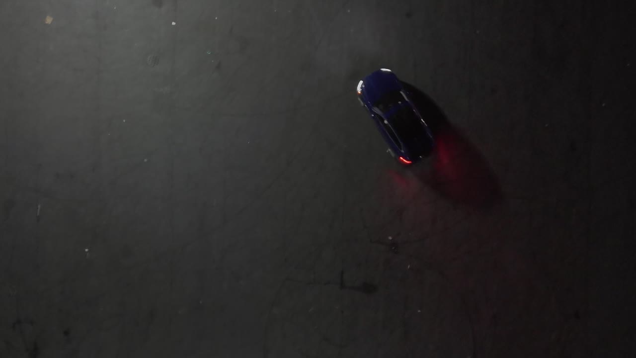 An aerial top down view of a blue SUV driving recklessly and doing doughnuts in an empty parking lot at night
