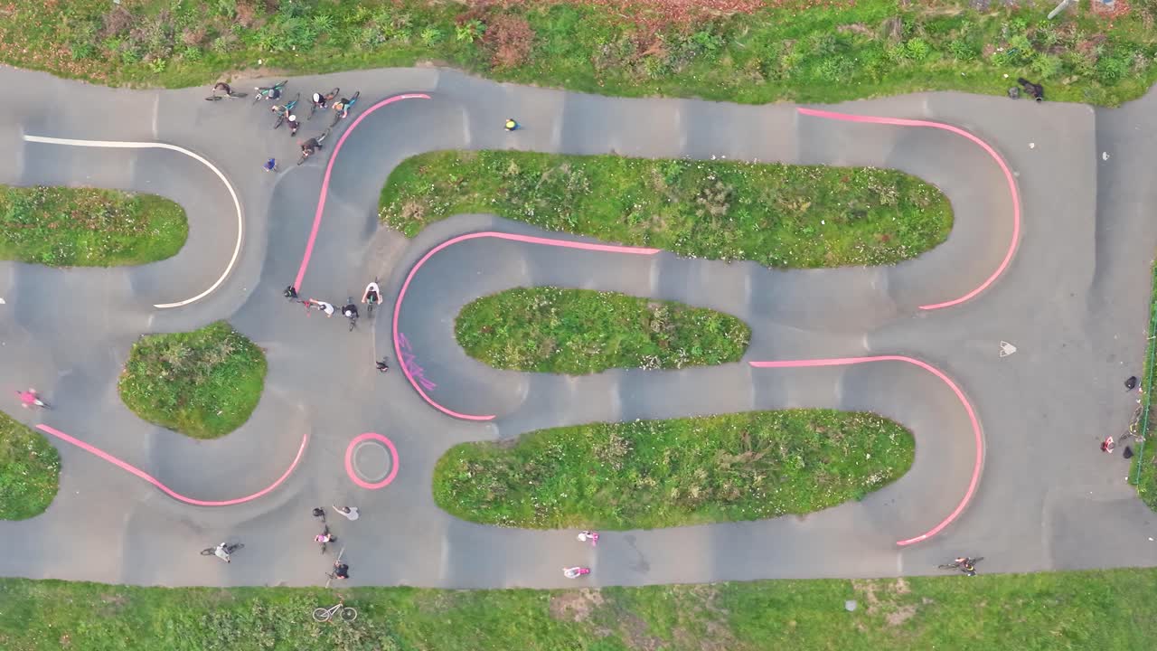 Top down static drone shot displays BMX freestyle riders flowing around the winding pump track in Hillsborough Park, Sheffield, South Yorkshire, England, highlighting curves, movement, and community