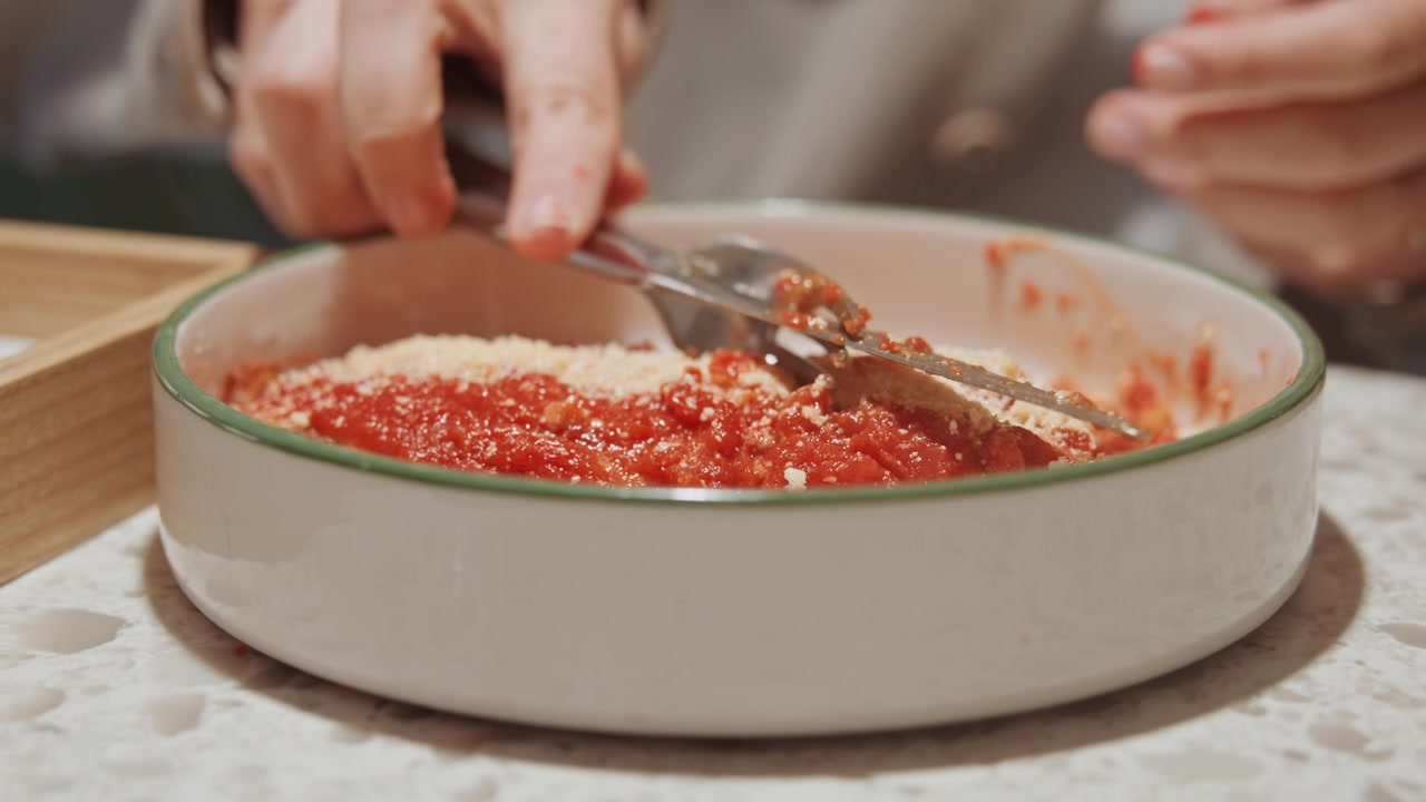 Primo piano di mani che mescolano salsa di pomodoro con formaggio grattugiato in una ciotola di ceramica bianca sul bancone. La persona usa due utensili di metallo per combinare pomodori rossi a pezzi e parmigiano giallo pallido, creando.
