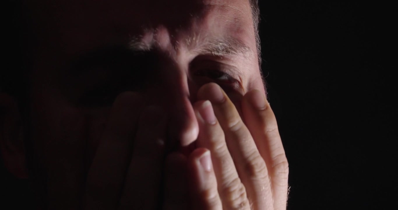 Close-up de um homem triste cobrindo o rosto com as mãos em um ambiente escuro, representando depressão, estresse, dor emocional e desafios de saúde mental