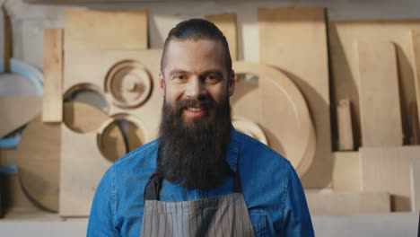 portrait of caucasian bearded man in apron looking at camera and smiling in carpentry workshop