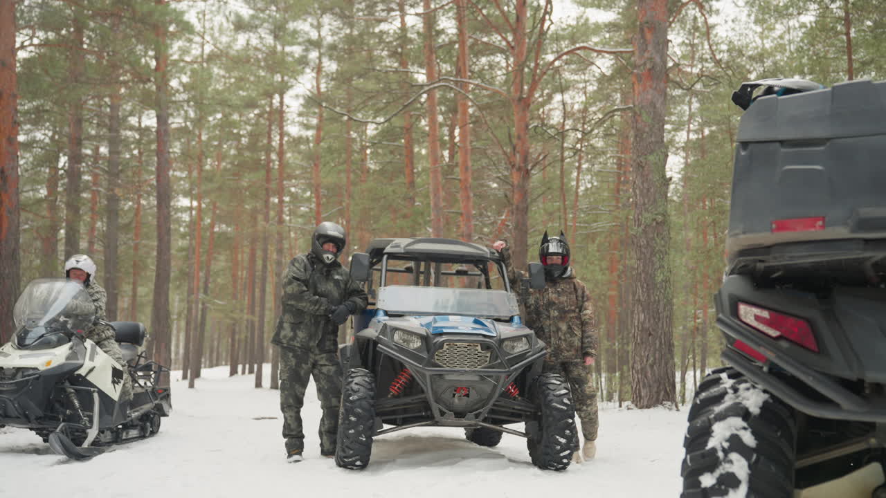 Un convoglio di ATV nella foresta innevata prepara i piloti con equipaggiamento mimetico. UTV side-by-side e motoslitte sono parcheggiati tra pini alti. Caschi indossati, motori al minimo, con il respiro visibile nel freddo. La squadra controlla il verricello e le sospensioni.