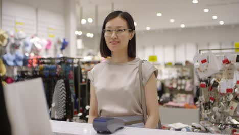 waist up shot of cheerful young woman pressing her smart watch to payment terminal at checkout counter of clothing store, taking shopping bags from sales assistant, thanking and leaving