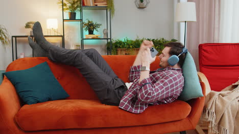 Man-in-wireless-headphones-listening-energetic-dancing-music-on-smartphone-relaxing-lying-on-sofa