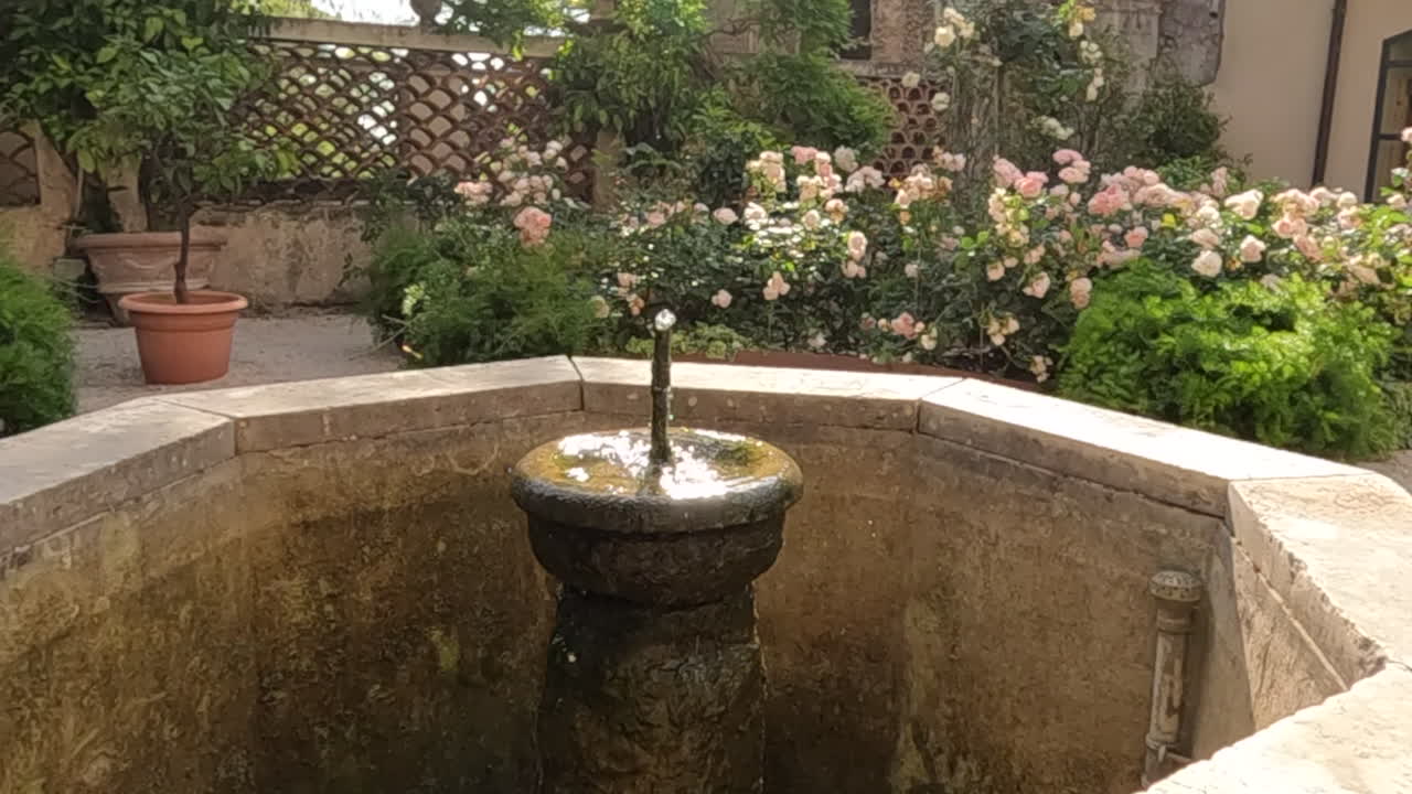 Encantadora fuente de piedra antigua fluye suavemente en un exuberante jardín italiano cerca de Tívoli, Roma. Rosas rosadas florecen junto a muros desgastados y macetas de terracota en este sereno patio histórico.